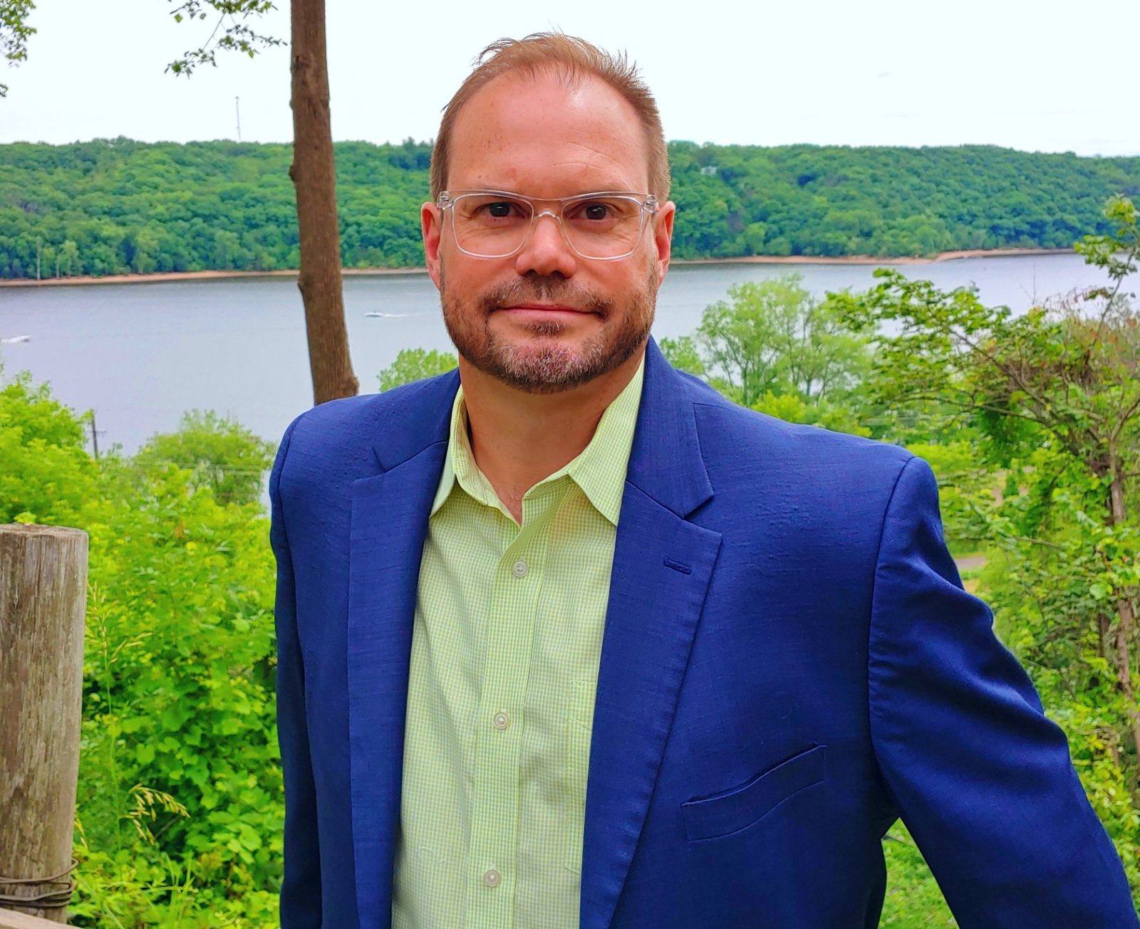 Photo of Darby Lunceford, a man in a light green shirt with a blue sports coat. Background is a scene with trees and a river.